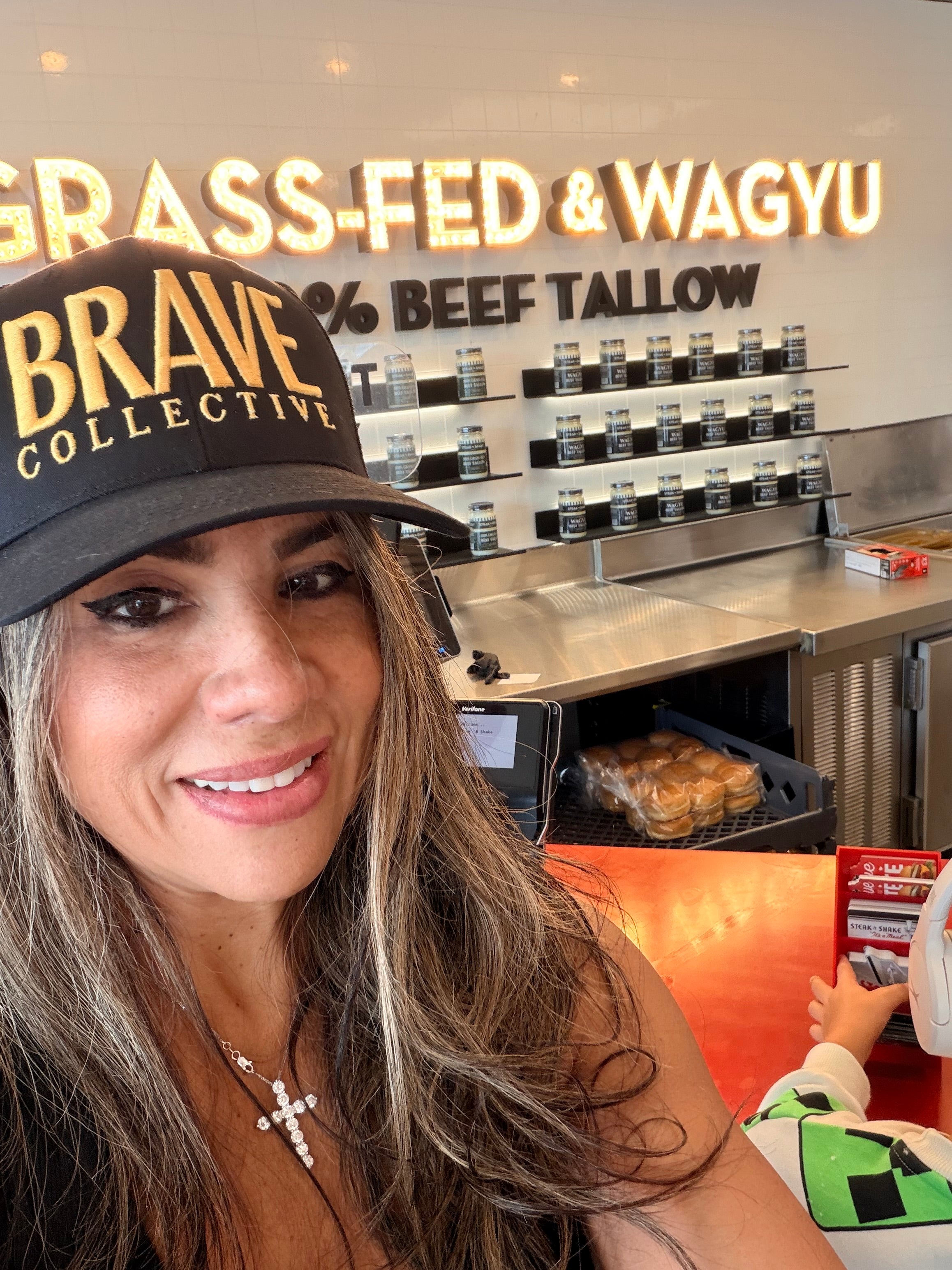 A woman in a restaurant setting wearing a black baseball cap with 'BRAVE COLLECTIVE' in gold lettering. 