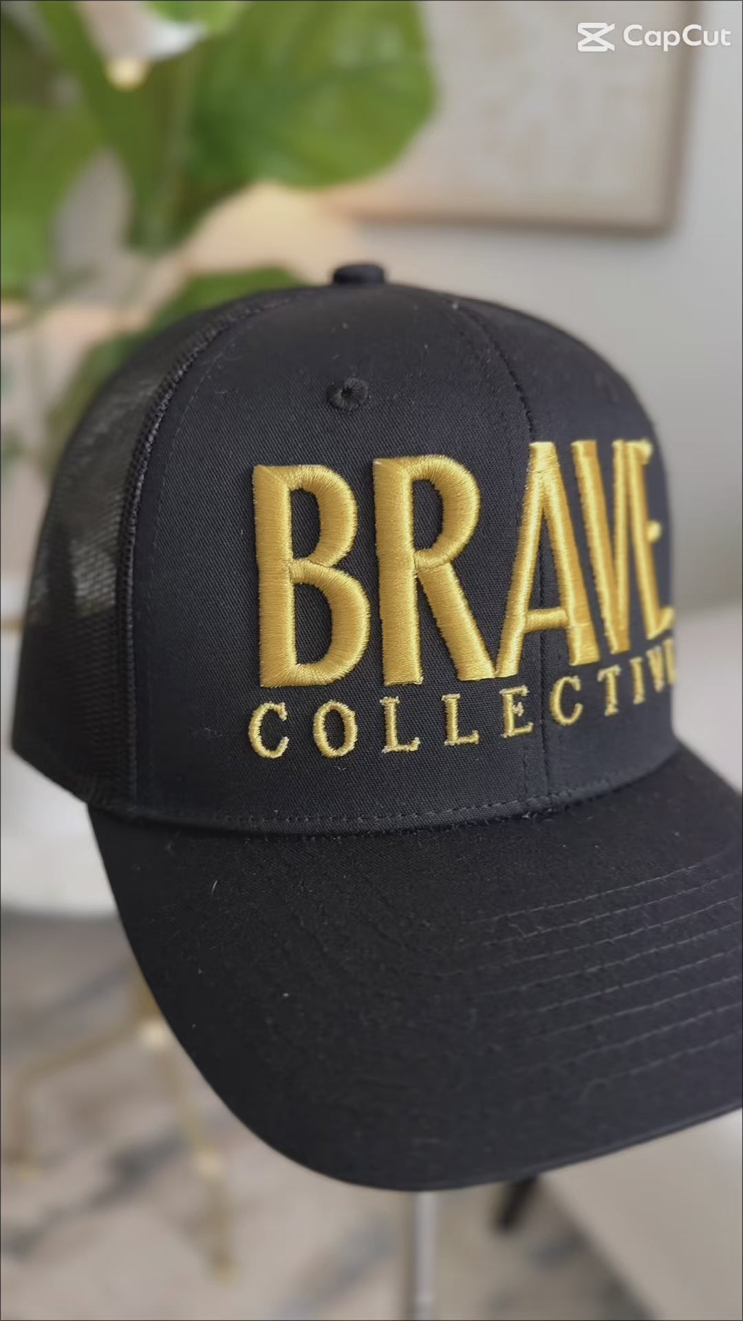 A black trucker-style baseball cap with gold embroidered text reading 'BRAVE COLLECTIVE' on the front, displayed indoors on a stand with a blurred plant and wall art in the background.