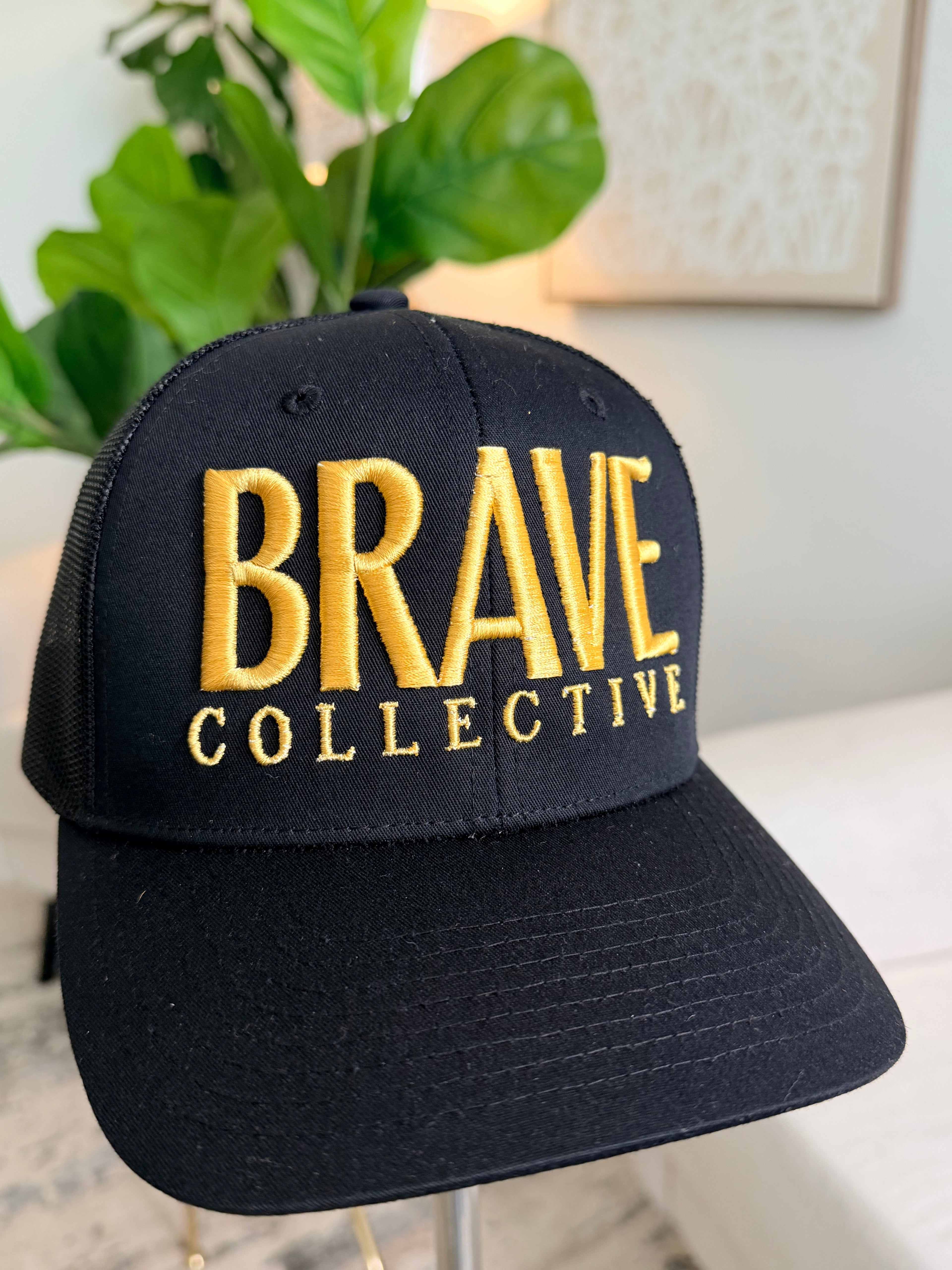 A black trucker-style baseball cap with gold embroidered text reading 'BRAVE COLLECTIVE' on the front, displayed indoors on a stand with a blurred plant and wall art in the background.